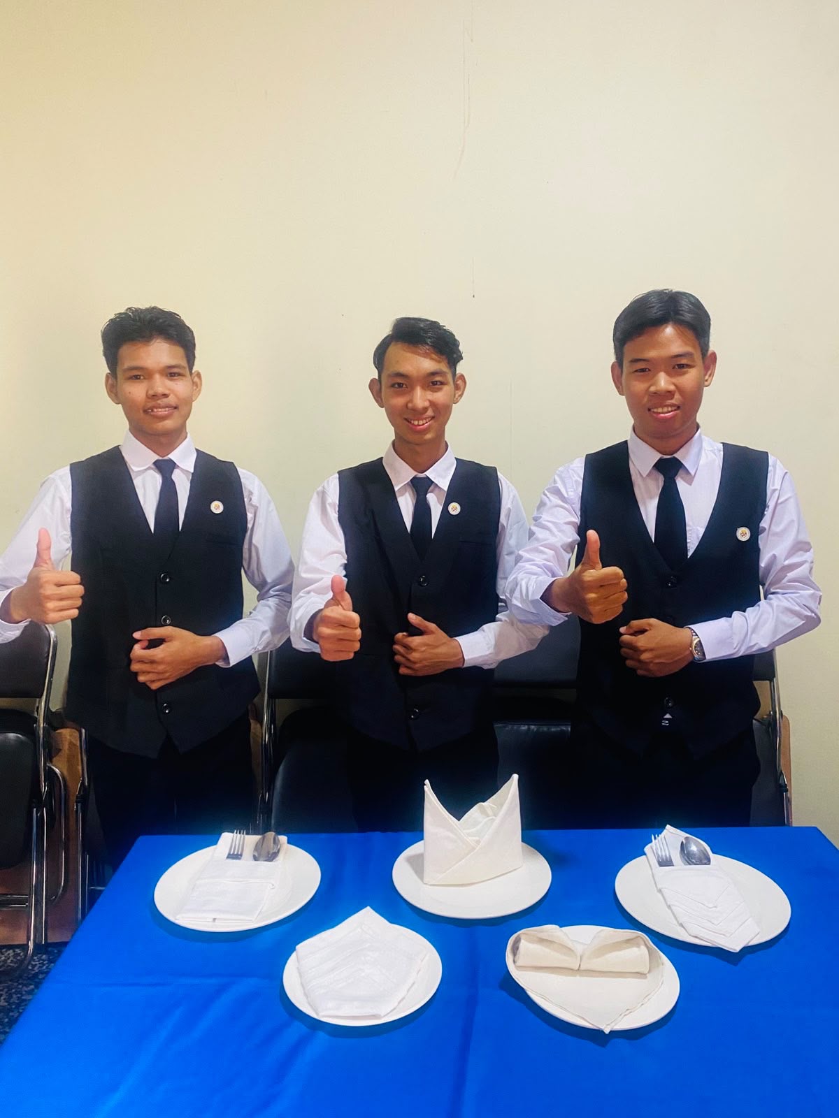 ပြင်ဆင်ထားသော စားပွဲတွင် ဝန်ဆောင်မှု လေ့ကျင့်နေသည့် HMTC သင်တန်းသား သုံးဦး